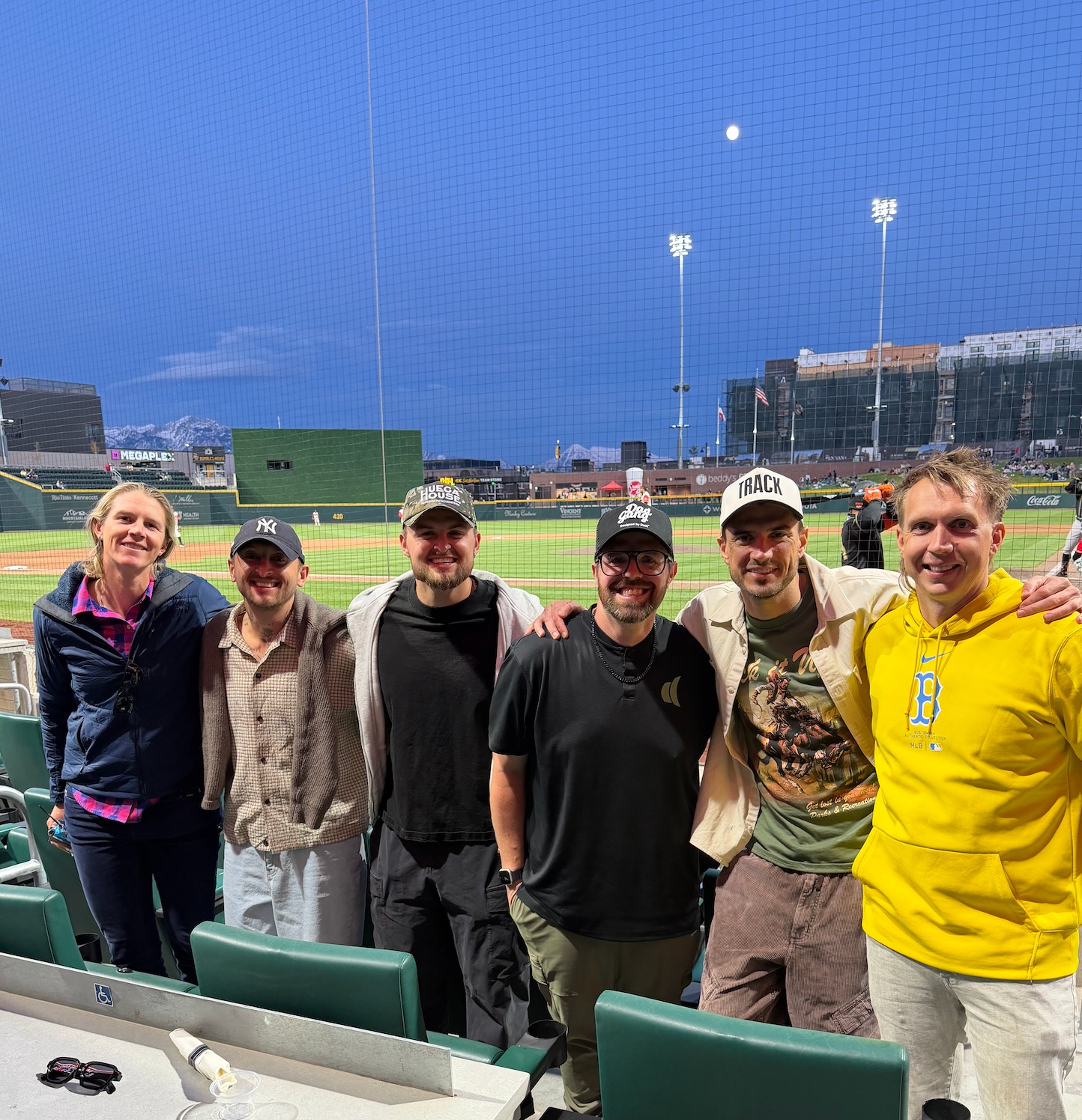 Six friends posing in green stadium seats at a ballpark with snow-capped mountains and a full moon behind
