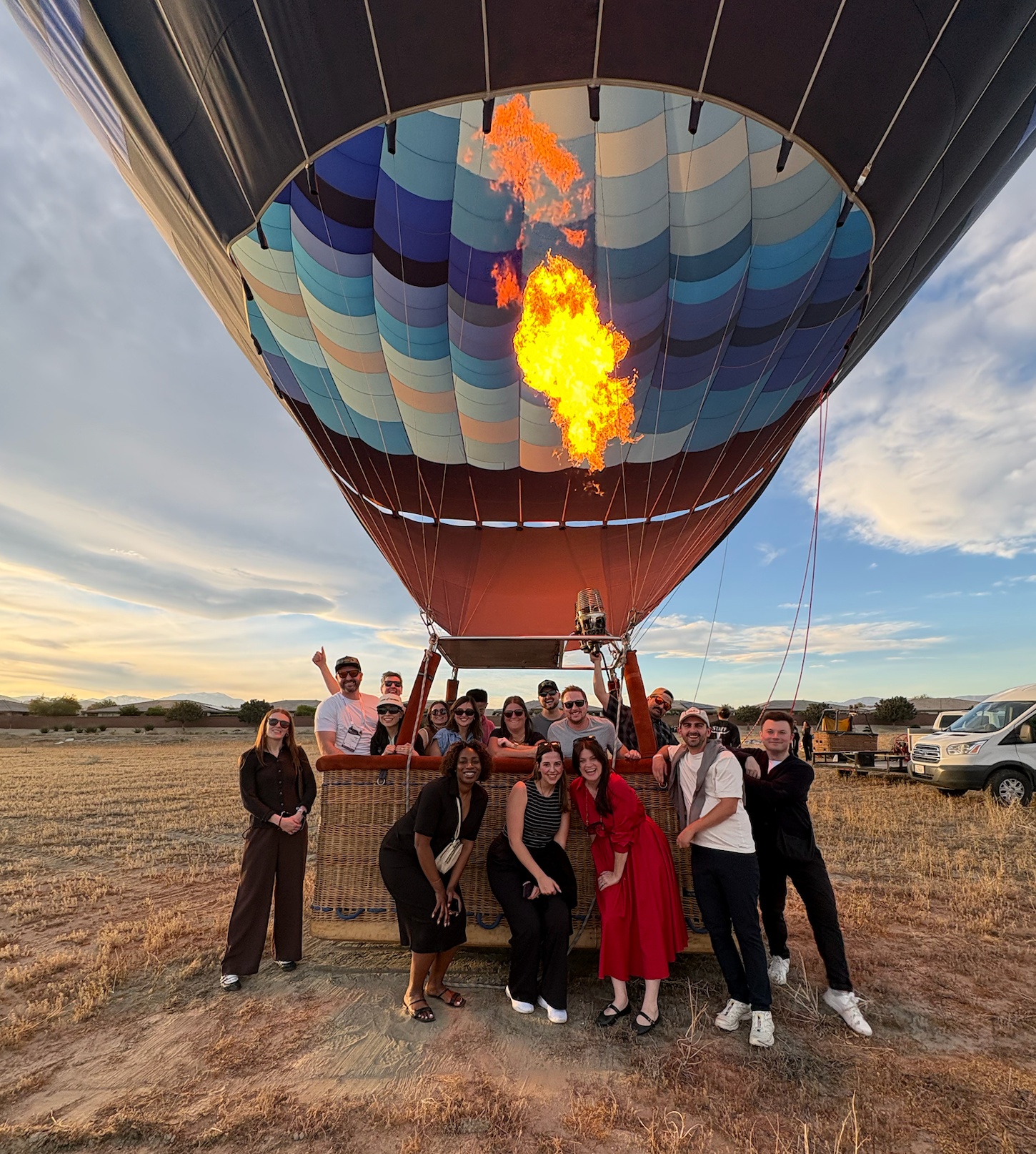 Large group posing under a colorful hot air balloon firing its flames in a field at golden hour